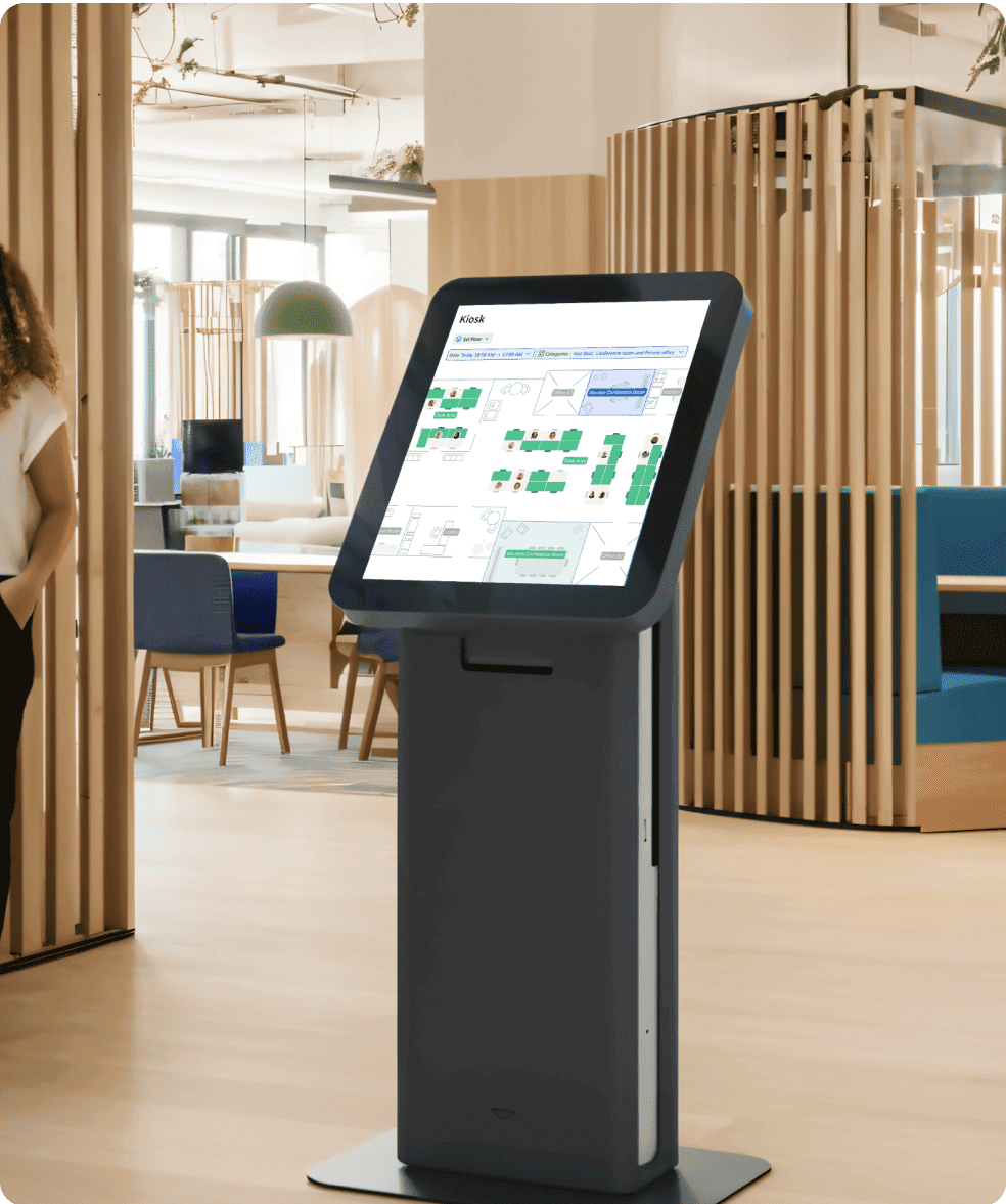 A kiosk screen in a coworking space displaying a floor plan for desk bookings with Archie app. The screen is mounted on a stand and shows available desks and office layout.
