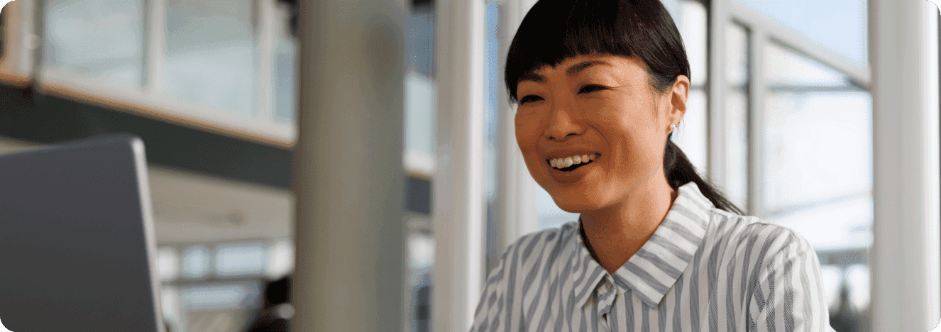 A smiling woman in a striped shirt working on a laptop in an office environment.