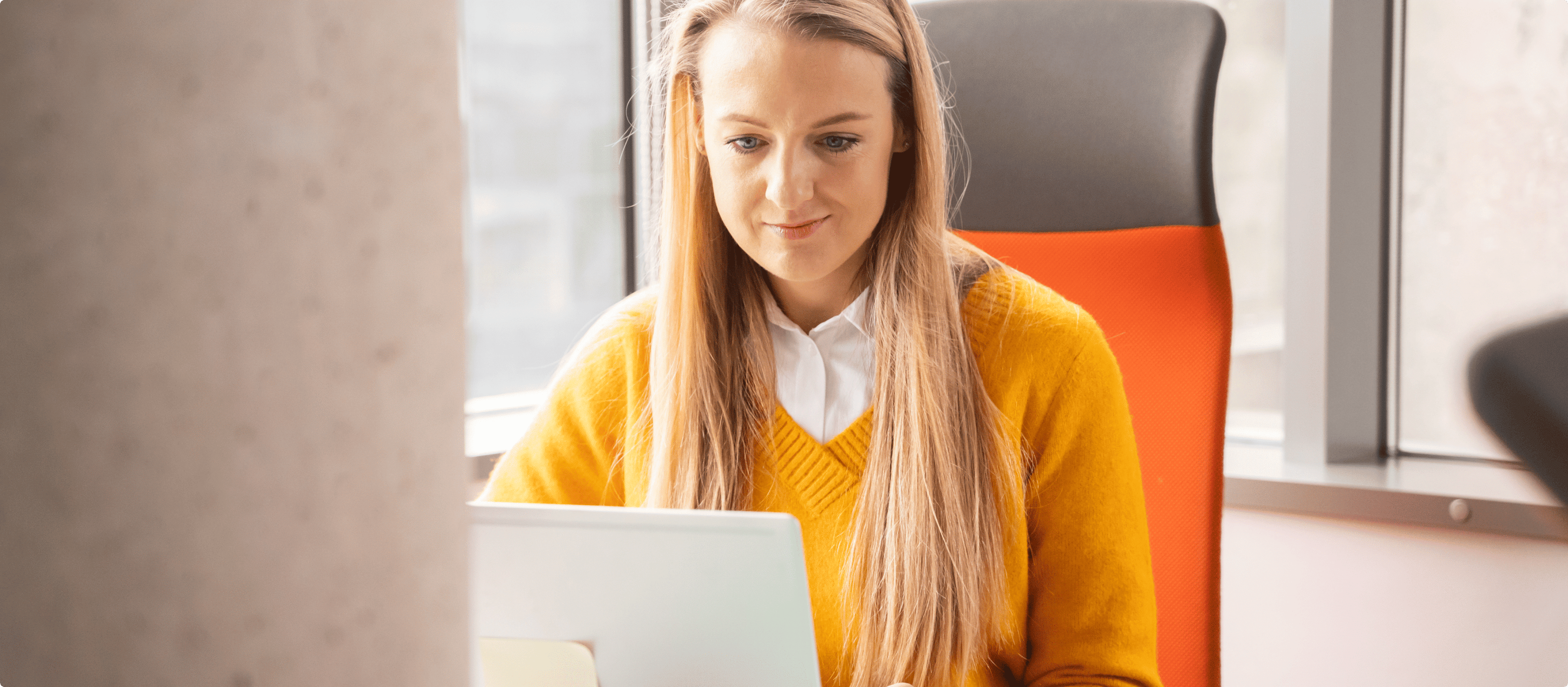 A woman with long blonde hair, wearing a yellow sweater, sitting at a desk in an office chair and working on a laptop.